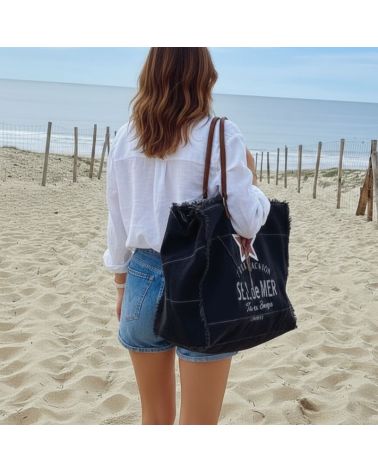 Femme de dos sur une plage de sable portant un grand sac cabas noir "Sel de Mer" au style bohème face à l'océan à Hossegor