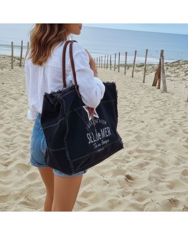 Grand sac de plage cabas noir en toile avec inscription "SEL de MER", franges et anses en cuir, porté à l'épaule sur une plage