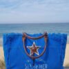 Sac de plage bleu roi XL Sel de Mer posé sur une rembarde en bois avec vue sur mer