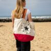 Sac de plage femme en toile écrue et rouge avec étoile noire, porté à l’épaule sur la plage face à l’océan.