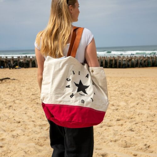Sac de plage femme en toile écrue et rouge avec étoile noire, porté à l’épaule sur la plage face à l’océan.