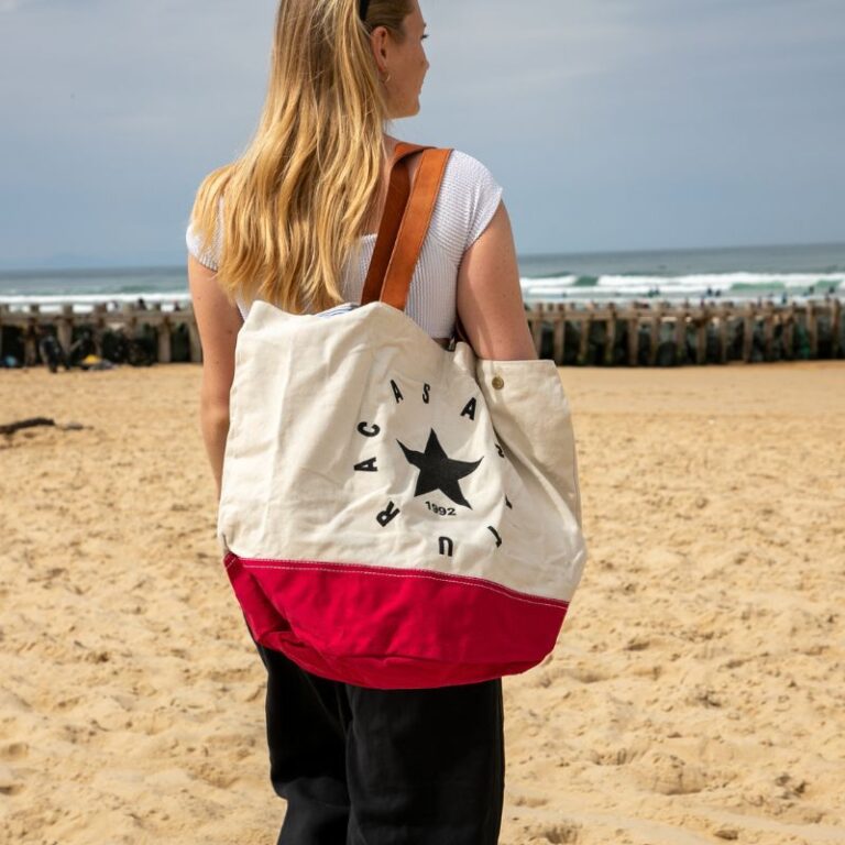 Sac de plage femme en toile écrue et rouge avec étoile noire, porté à l’épaule sur la plage face à l’océan.